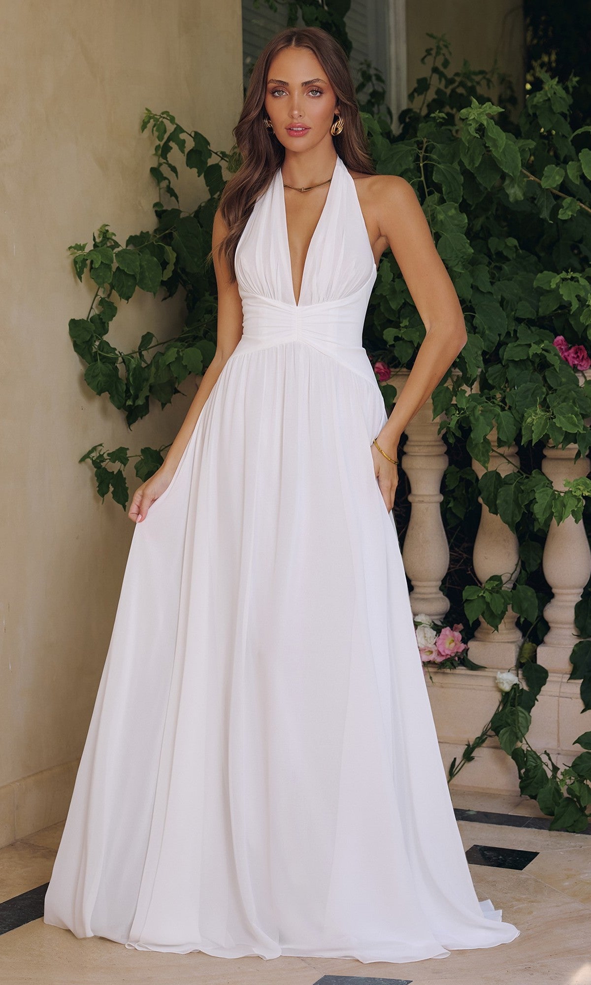 A woman in the Amoris T715W long off-white halter prom dress stands outdoors before lush green foliage and a stone railing.